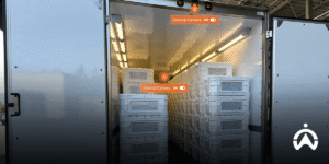The warehouse door opens to reveal a stack of logistics crates with a camera surveillance system.