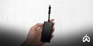 Tangan memegang perangkat GPS tracker hitam modern dengan kabel di depan dinding putih untuk sistem pelacakan kendaraan.
