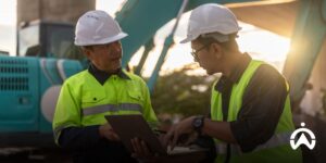Dua pekerja konstruksi meninjau data operasional di laptop untuk pangkas biaya armada perusahaan menggunakan sistem telematika di lokasi proyek.