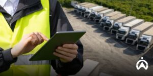 Petugas dengan rompi keselamatan menggunakan tablet untuk kustomisasi fleet management system dalam memantau operasional truk di area parkir.