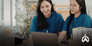 Two women in blue work together on a laptop next to a donation box.