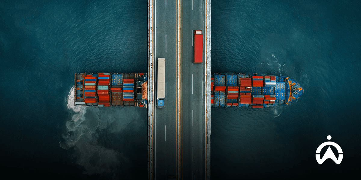 Foto udara jembatan dengan dua truk melintas dan dua kapal kontainer di bawahnya, menggambarkan sistem manajemen armada dalam logistik multi-moda