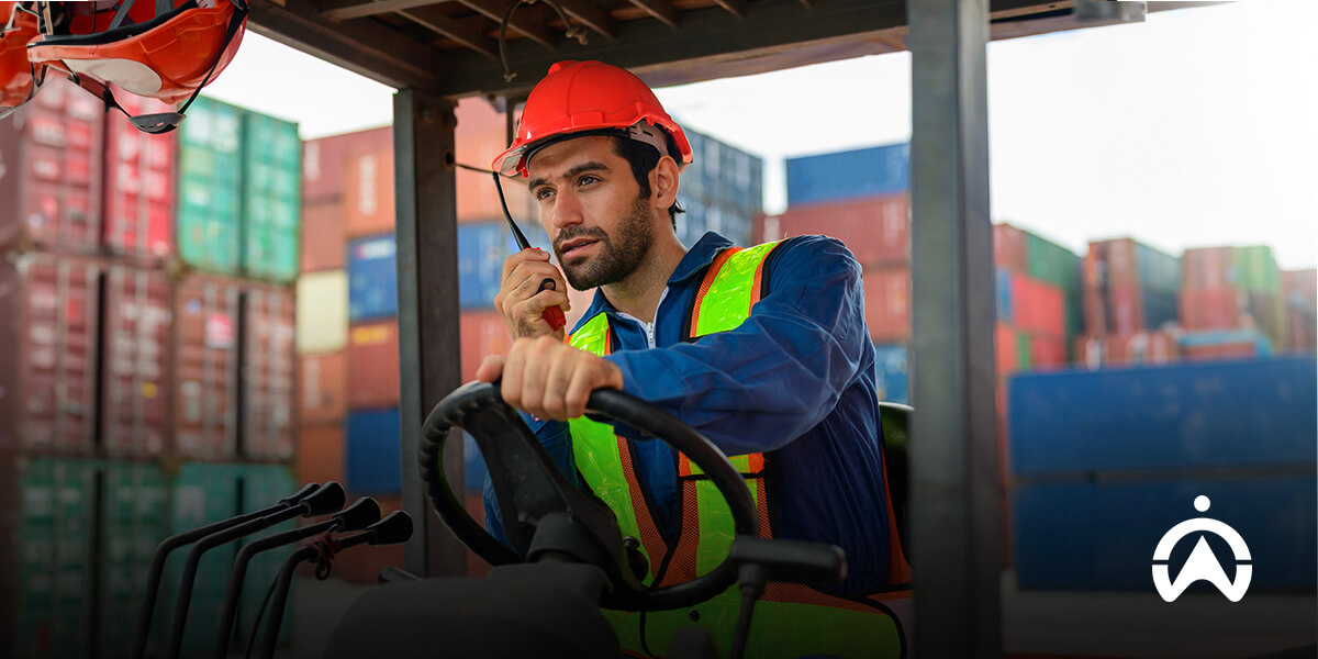 Pekerja dengan alat keselamatan mengoperasikan forklift di depo kontainer, mencerminkan salah satu tugas utama manajer armada.