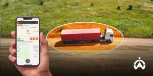 Tangan memegang smartphone dengan aplikasi sistem pemantauan kendaraan logistik, berlatar belakang truk merah di jalan.