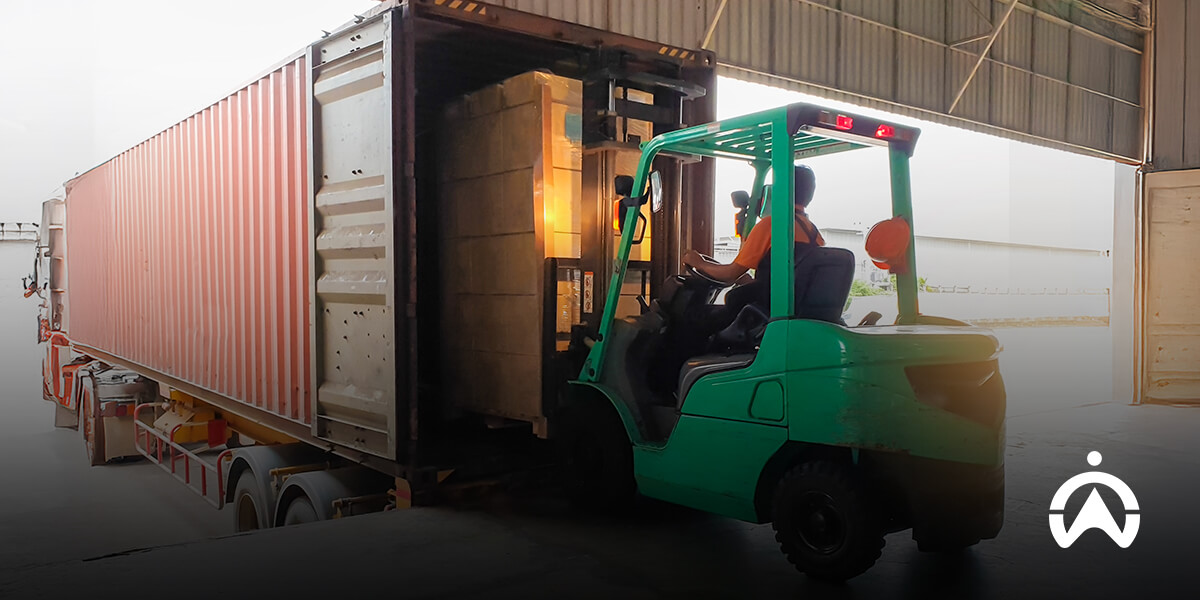 A green forklift operator is loading large cardboard boxes into a container for large-scale shipping.