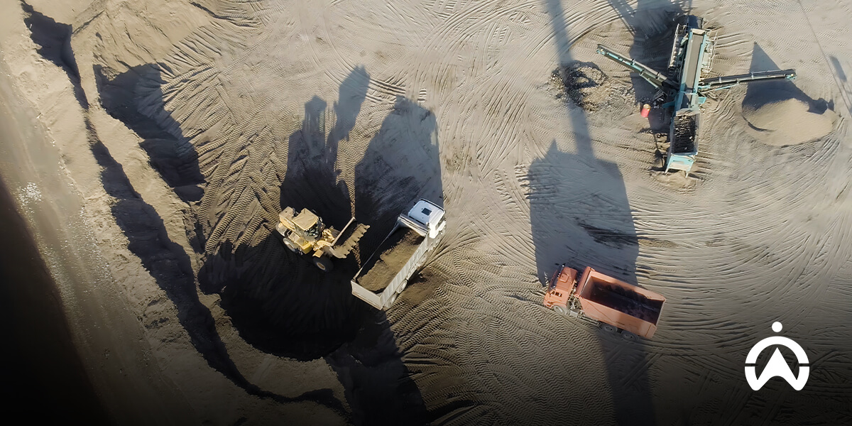 An aerial photo of a yellow excavator loading sand into a truck, showing one type of heavy equipment based on its function in the mining area.